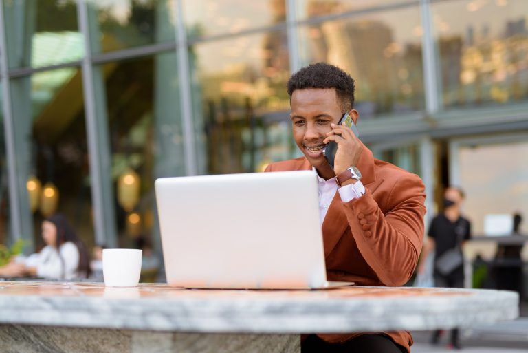 Happy African businessman outdoors in coffee shop using laptop computer while talking on mobile phone